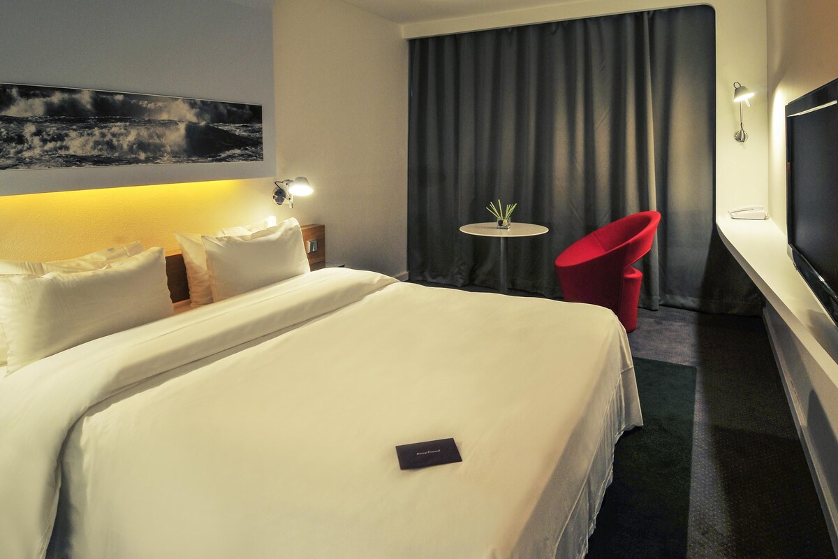 Hotelkamer met een groot bed, een klein tafeltje met een vaas, een rode stoel en een televisie aan de muur.