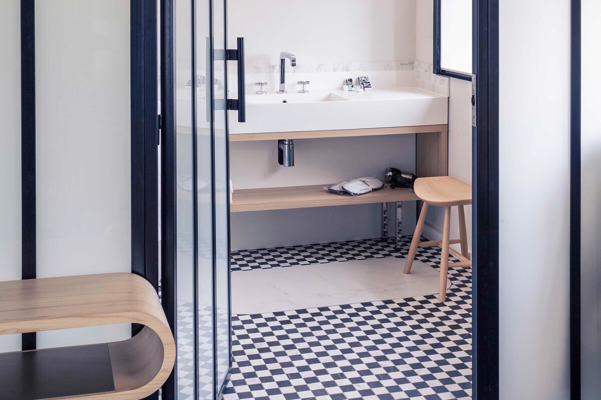 Une salle de bain moderne avec un carrelage à motifs géométriques, un banc en bois et un lavabo blanc surmonté d’un miroir.