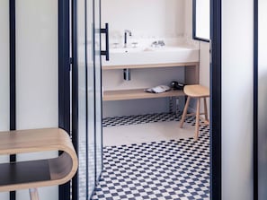 Une salle de bain moderne avec un carrelage à motifs géométriques, un banc en bois et un lavabo blanc surmonté d’un miroir.