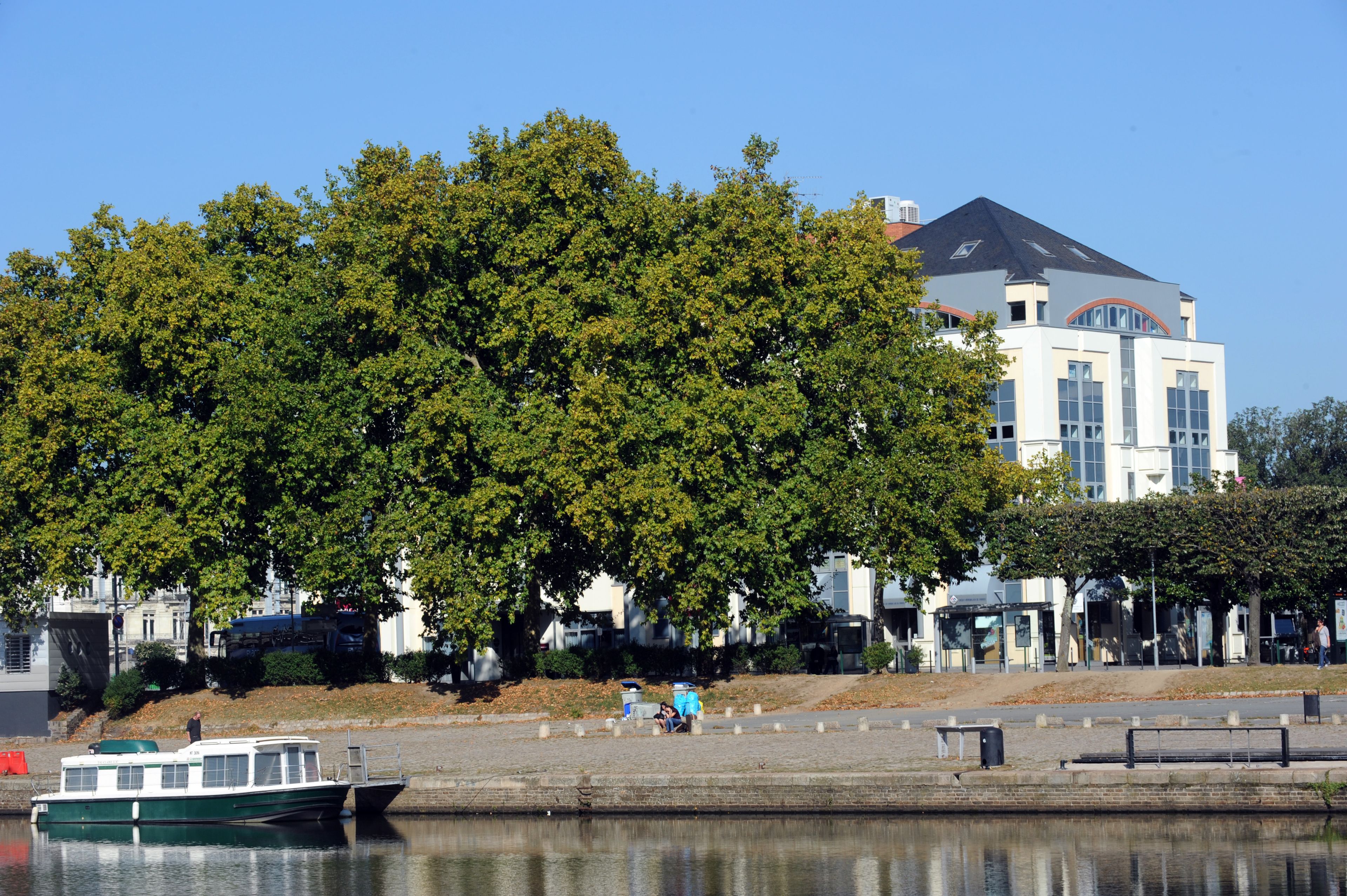 Ein Fluss mit einem vertäuten Boot am linken Ufer, ein großer Baum im Vordergrund und ein Gebäude mit geneigtem Dach im Hintergrund.