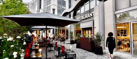 Un área de comedor al aire libre con mesas y sillas, un paraguas grande y un edificio con letrero de restaurante.