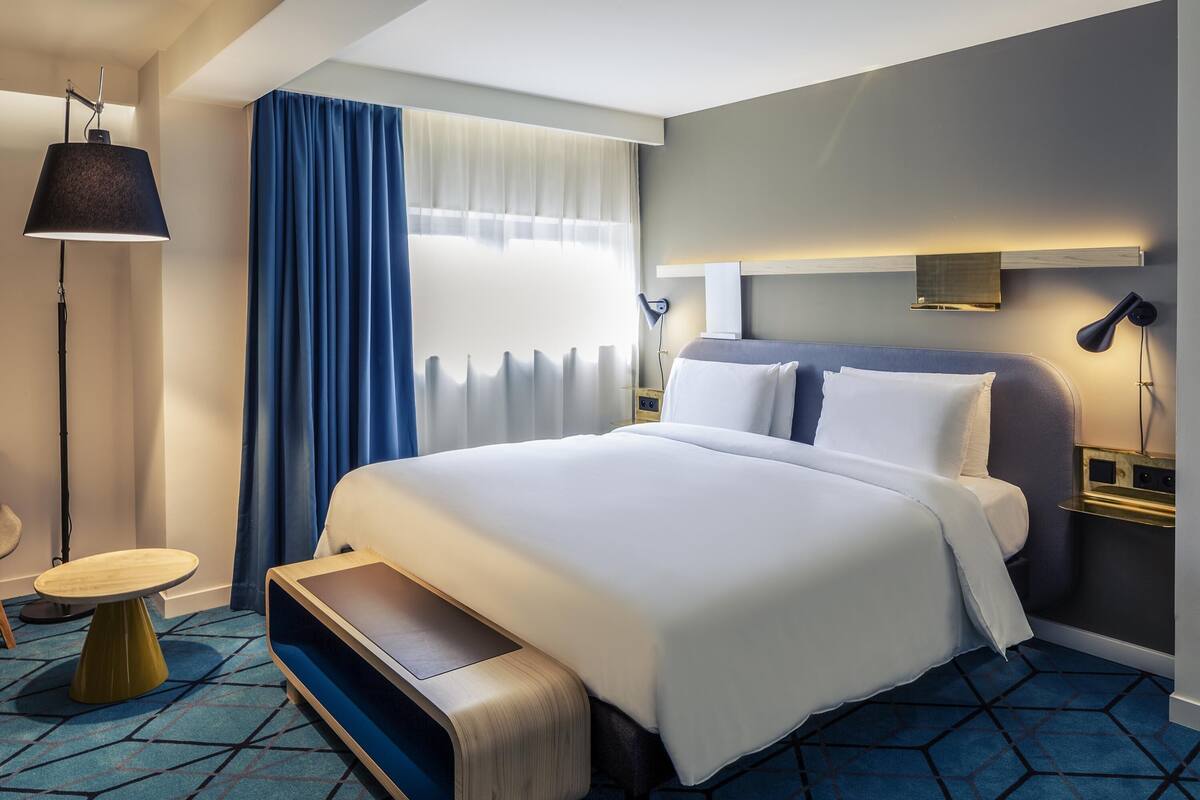 A modern hotel room with a large bed, a desk, a chair, and a lamp.