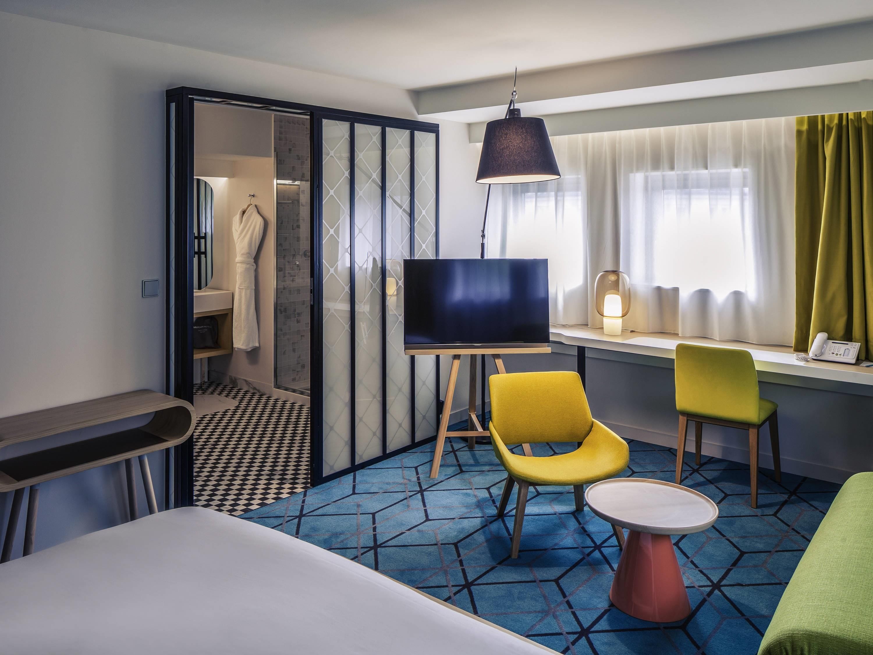 Et hotelværelse med en seng, et fjernsyn på et stativ, to stole, et lille bord, et bruseområde og et vindue med gardiner.
