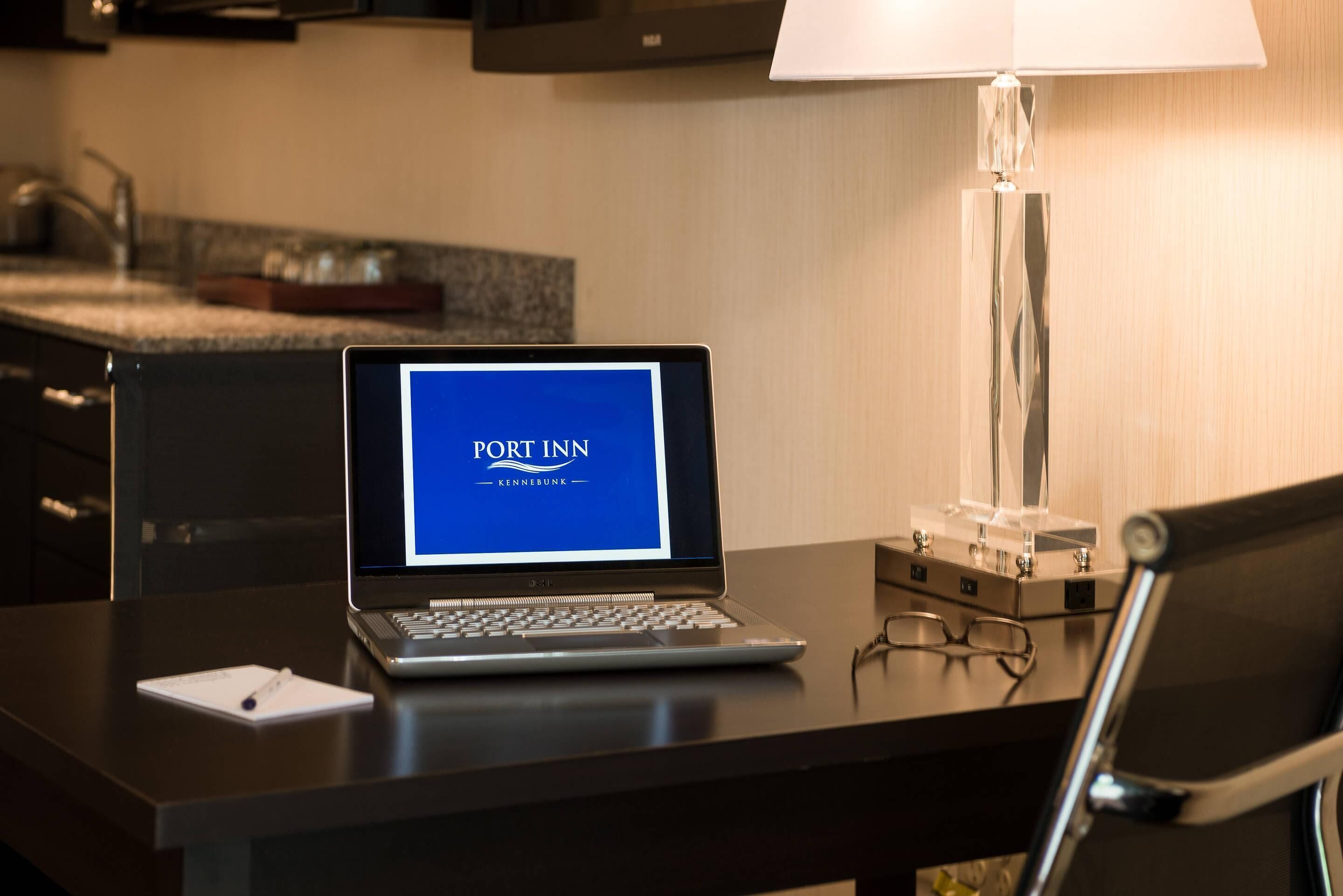 Ein Laptop auf einem Schreibtisch, mit einem 'PORT INN' Bildschirm, einer Lampe und einer Brille.