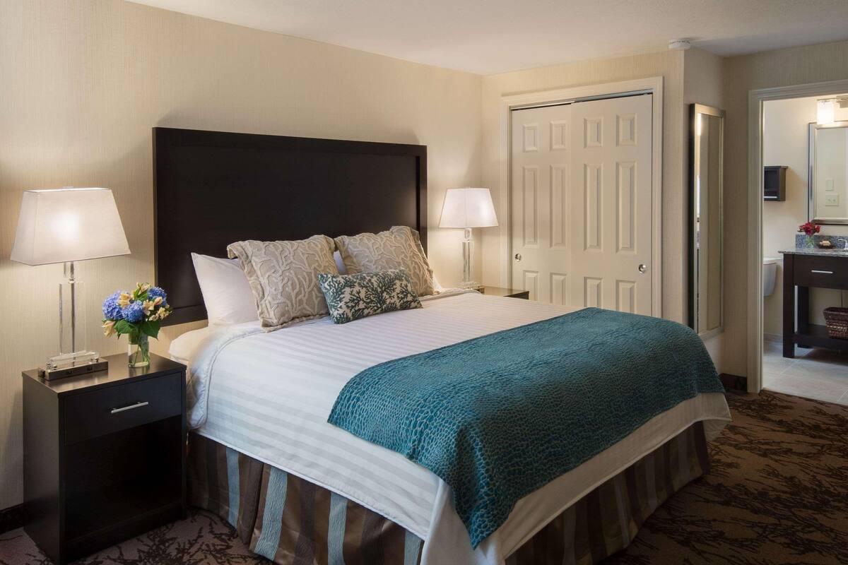 A hotel room with a bed, two bedside lamps, a nightstand, a dresser, and a door.