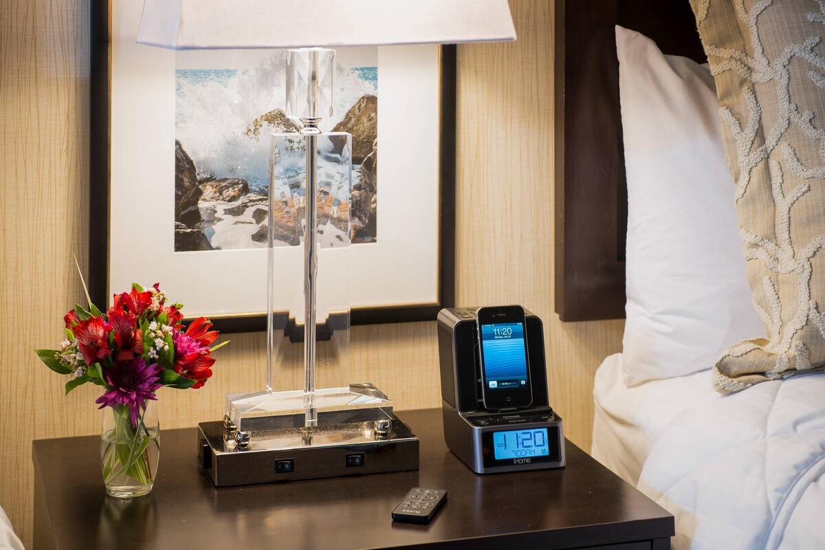 A hotel room with a bedside table featuring a lamp, a clock, a remote control, and a vase with flowers.