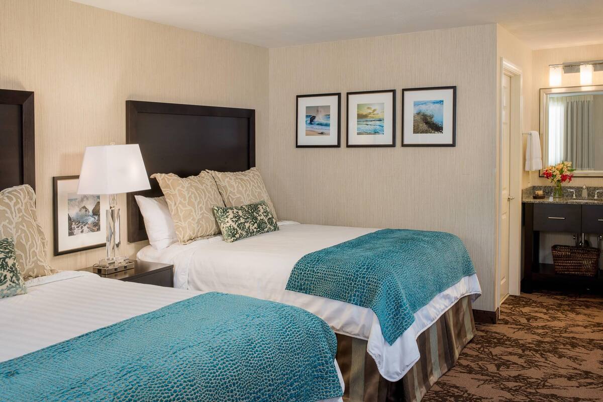 A hotel room with two beds, a nightstand with a lamp, a framed picture, and a bathroom visible through an open door.