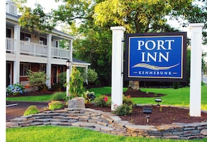 A sign for Port Inn Kennebunk, a two-story building with a balcony, surrounded by well-maintained landscaping.