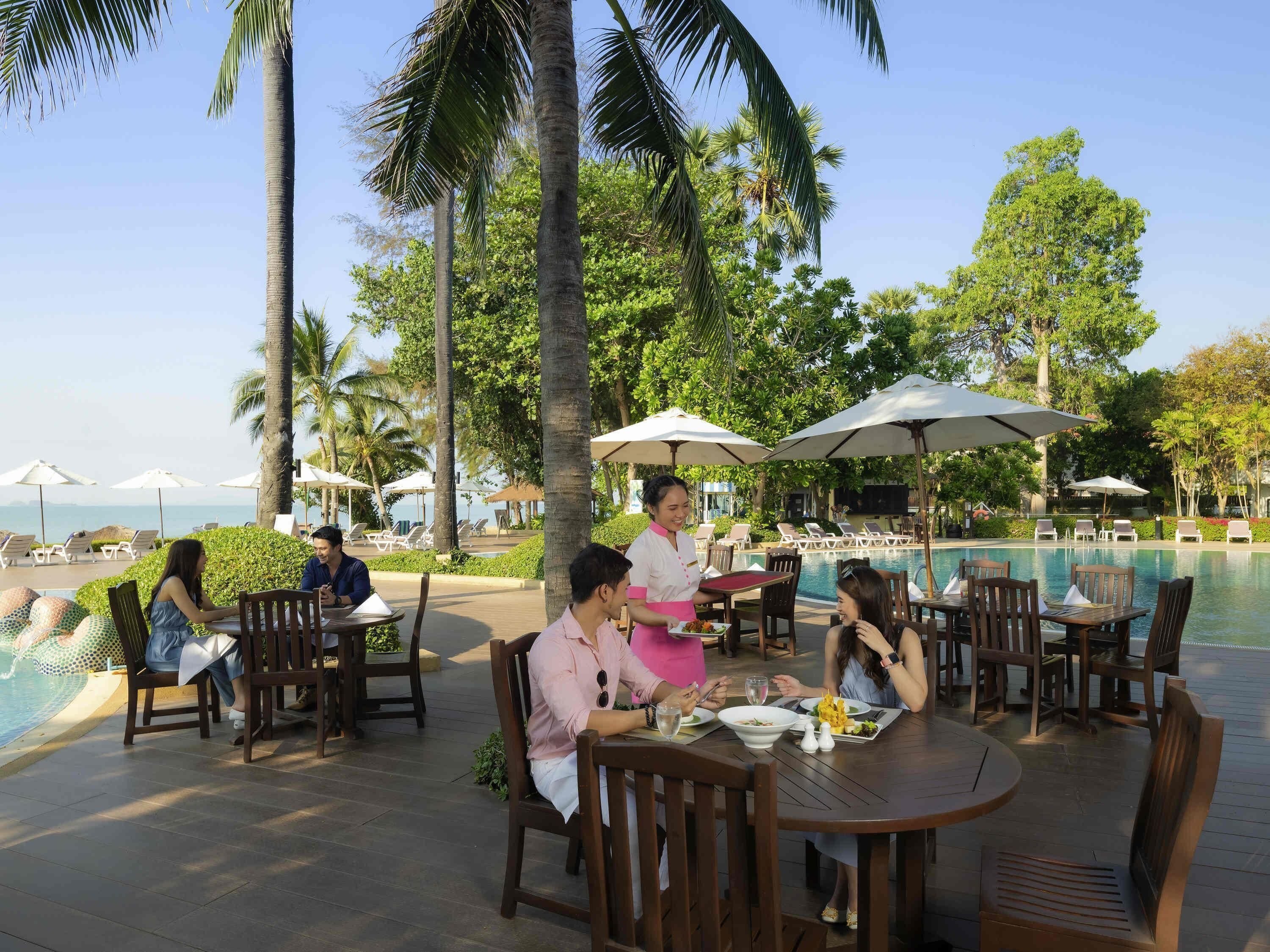 A poolside dining area with tables and chairs, palm trees, and a view of the sea.