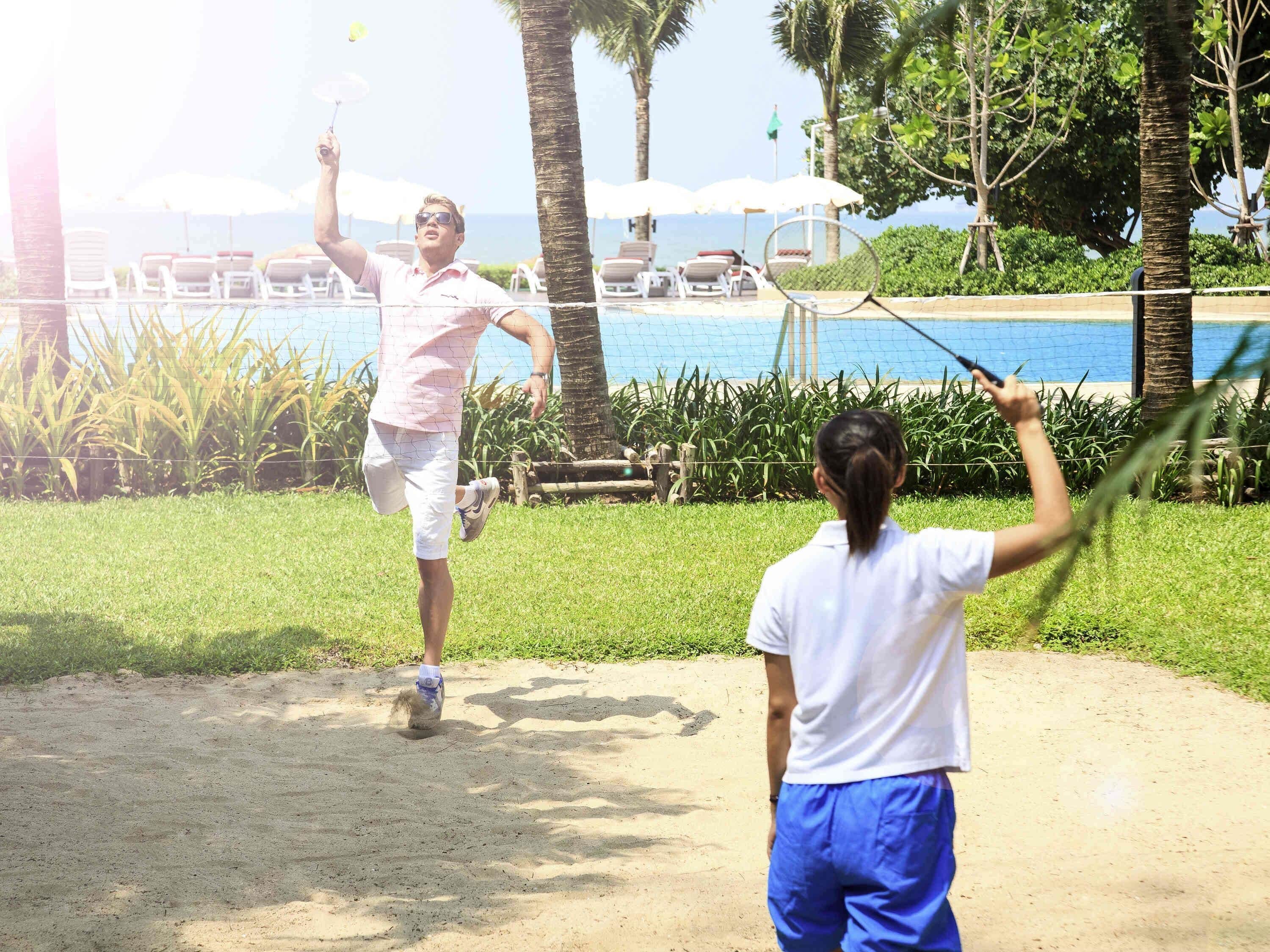 A man and a woman playing badminton in a sunny outdoor area with a pool and lounge chairs in the background.