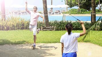 A man and a woman playing badminton in a sunny outdoor area with a pool and lounge chairs in the background.