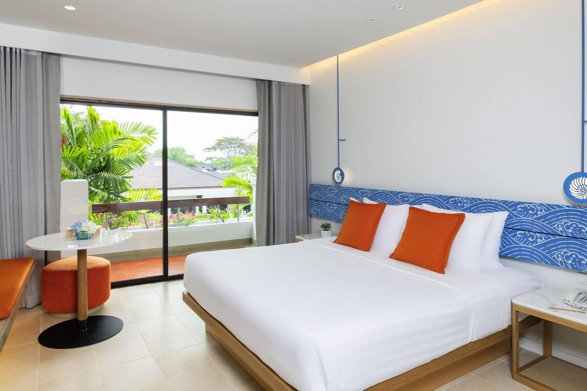 Habitación de hotel con cama, una mesita, una silla, ventana con vistas a zonas verdes y una lámpara en el techo.