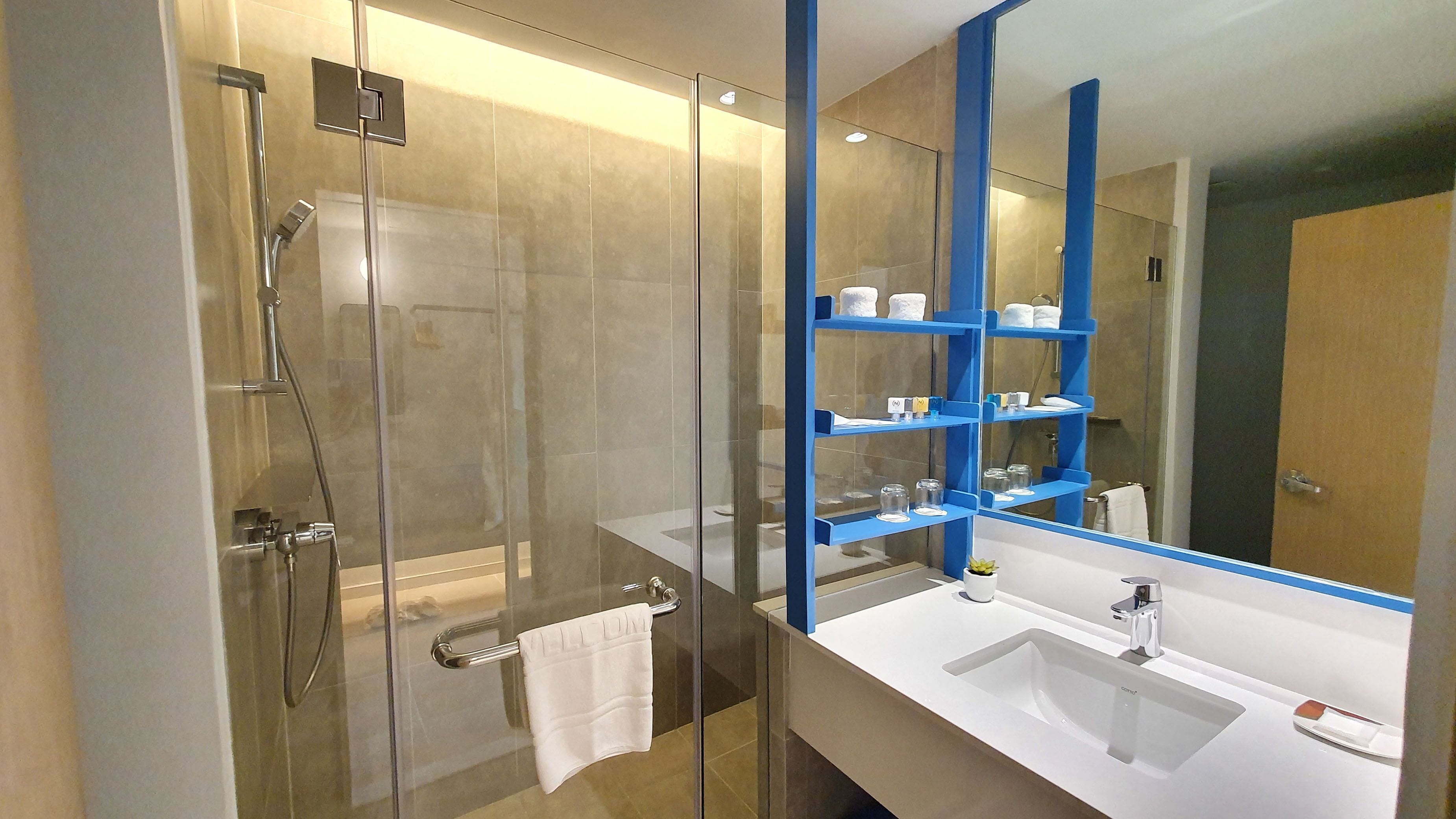 A modern bathroom with a glass-enclosed shower, white sinks, and a blue shelf unit with toiletries.