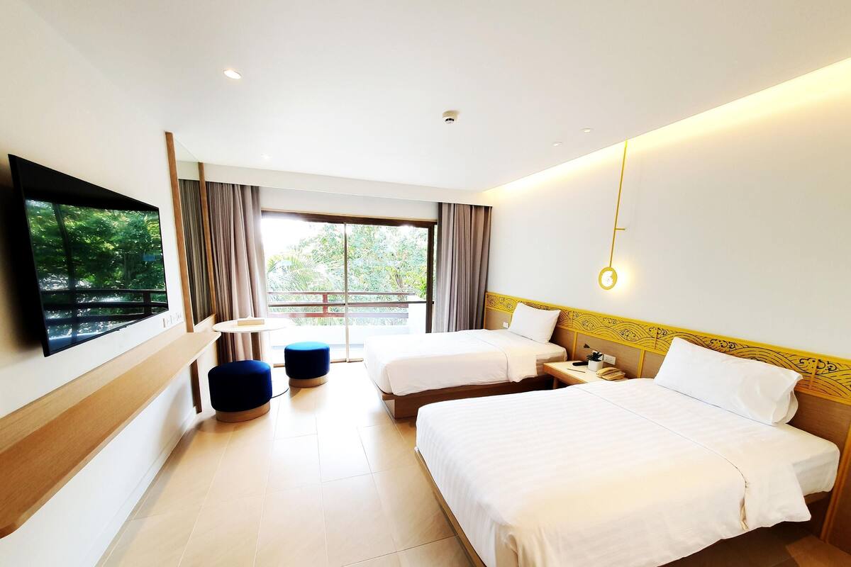 Habitación de hotel con dos camas, televisión, una mesita y balcón con vistas a zonas verdes.