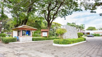 A paved entrance with a small kiosk, a stone wall, and a large tree.