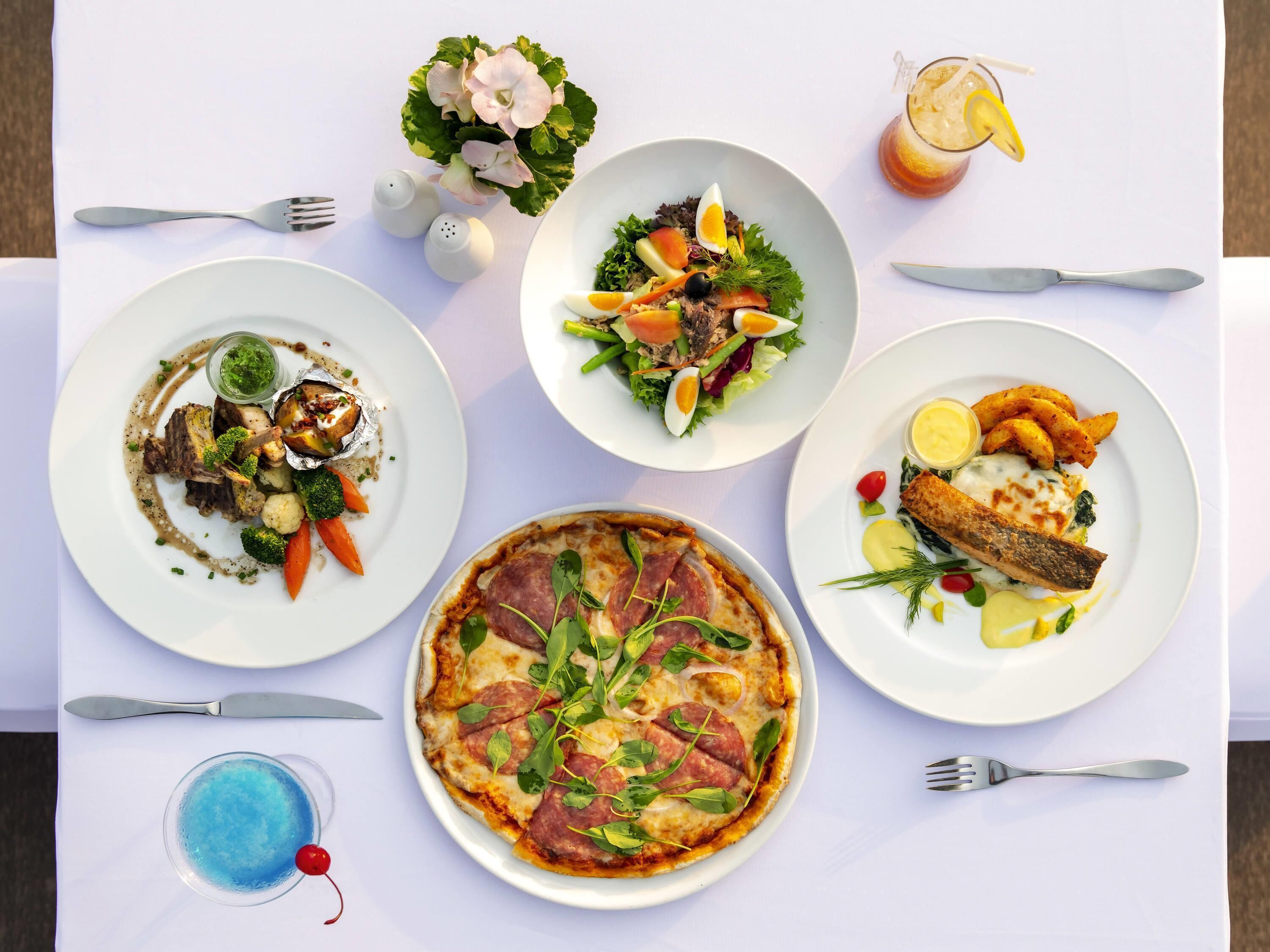 A table set with various dishes including a pizza, salad, and a steak dish, along with drinks and cutlery.