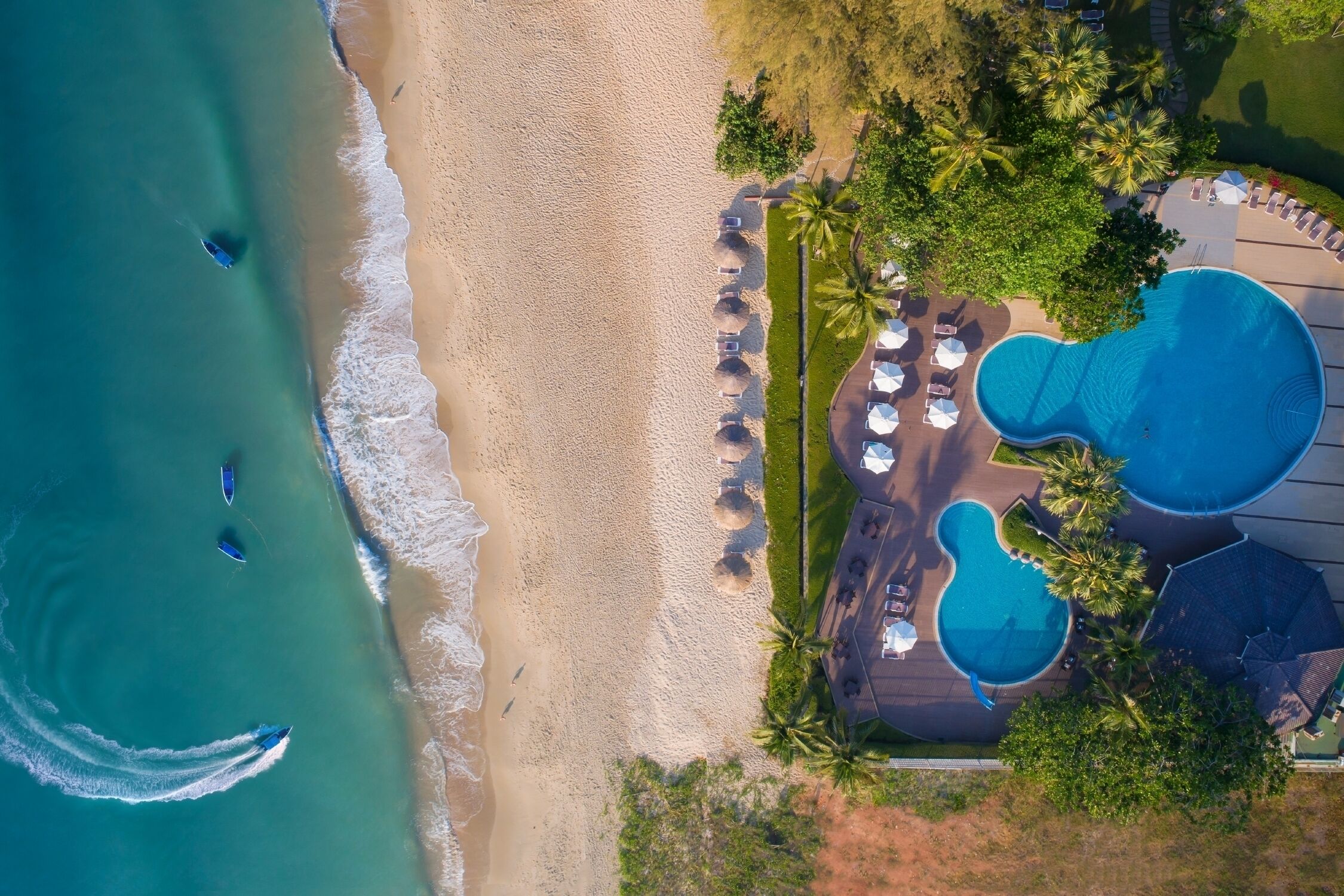 An aerial view of a beach resort with a swimming pool area, beach umbrellas, and palm trees.