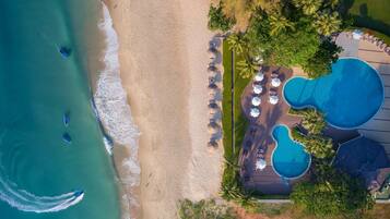 An aerial view of a beach resort with a swimming pool area, beach umbrellas, and palm trees.