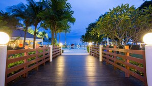 A wooden pathway leading to a pool area, flanked by palm trees and illuminated by lights. - Novotel Rayong Rim Pae Resort (Klaeng)