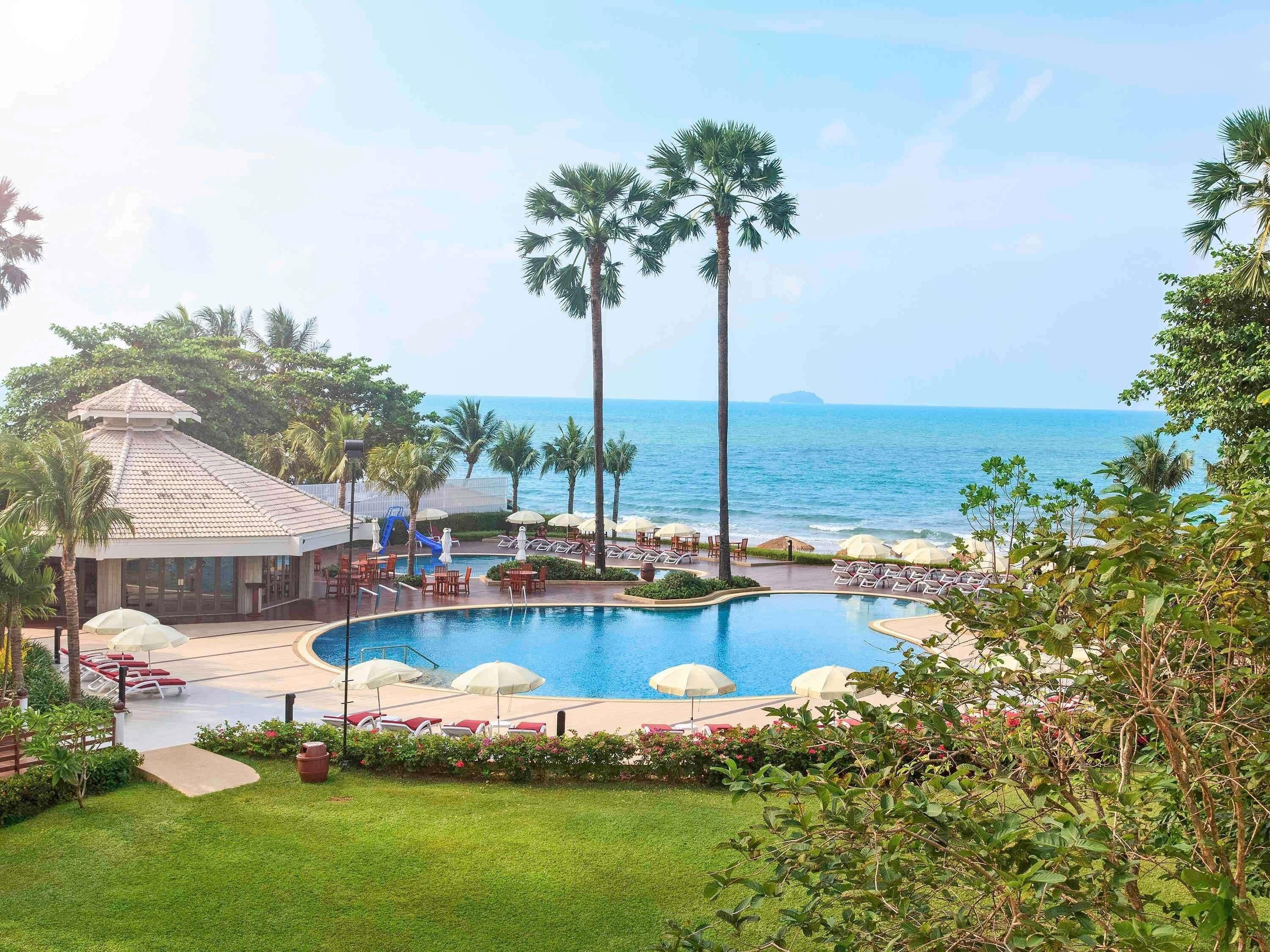 A resort pool with a view of the ocean, surrounded by palm trees and lounge chairs.