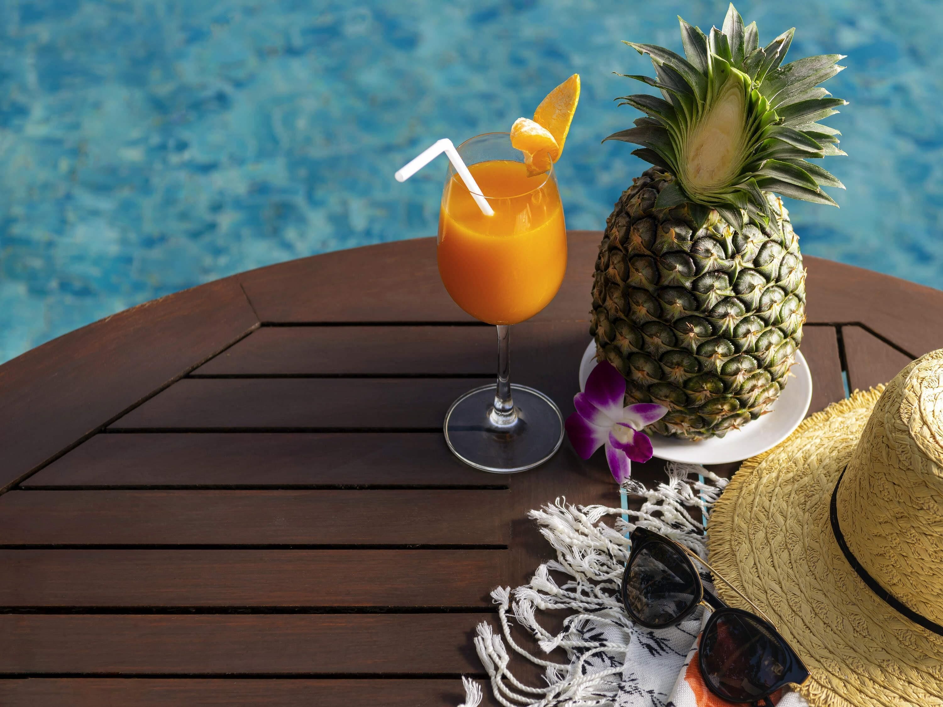 Cenário tropical com mesa de madeira junto a uma piscina, apresentando uma abacaxi, uma bebida com canudo, óculos de sol e um chapéu de palha.