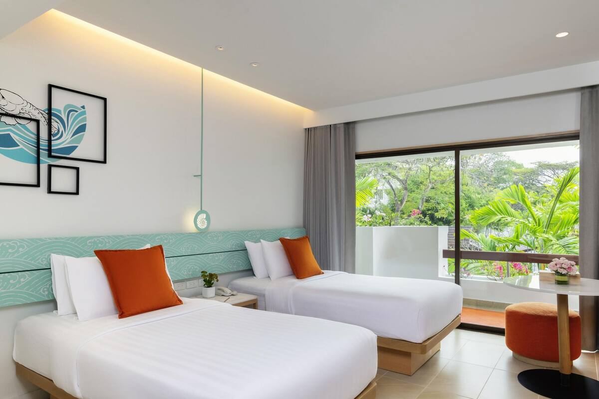 Habitación de hotel con dos camas, una mesita con un florero y un balcón con vistas a zonas verdes.