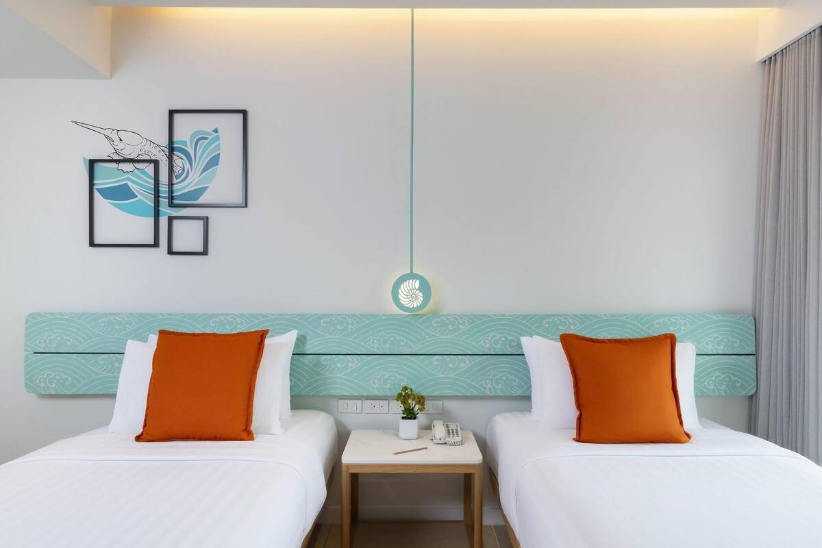 Habitación de hotel con dos camas, una mesita de noche, una lámpara fijada a la pared y obras de arte enmarcadas.