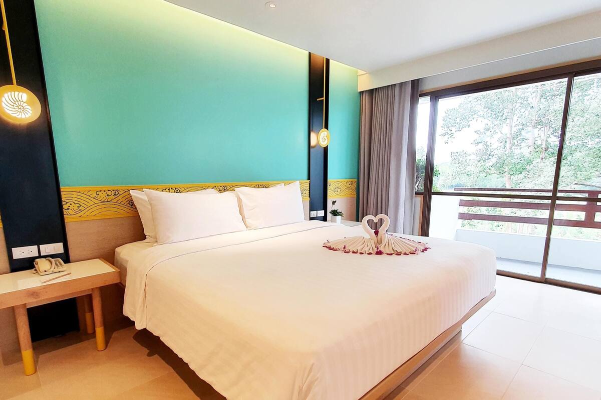 Habitación de hotel con cama, mesita de noche y vistas a la vegetación a través de amplias ventanas.