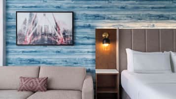 Ein Hotelzimmer mit Bett, Nachttisch, einem Stadtbild in einem Bilderrahmen und einer Couch mit einem gemusterten Kissen.