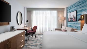 Una habitación de hotel con una cama grande, un escritorio con silla, televisor, espejo, lámpara y un cuadro en la pared.