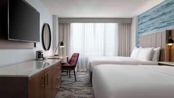 A hotel room with a flat-screen TV, a desk with a lamp, a chair, two beds, and a large window with curtains.