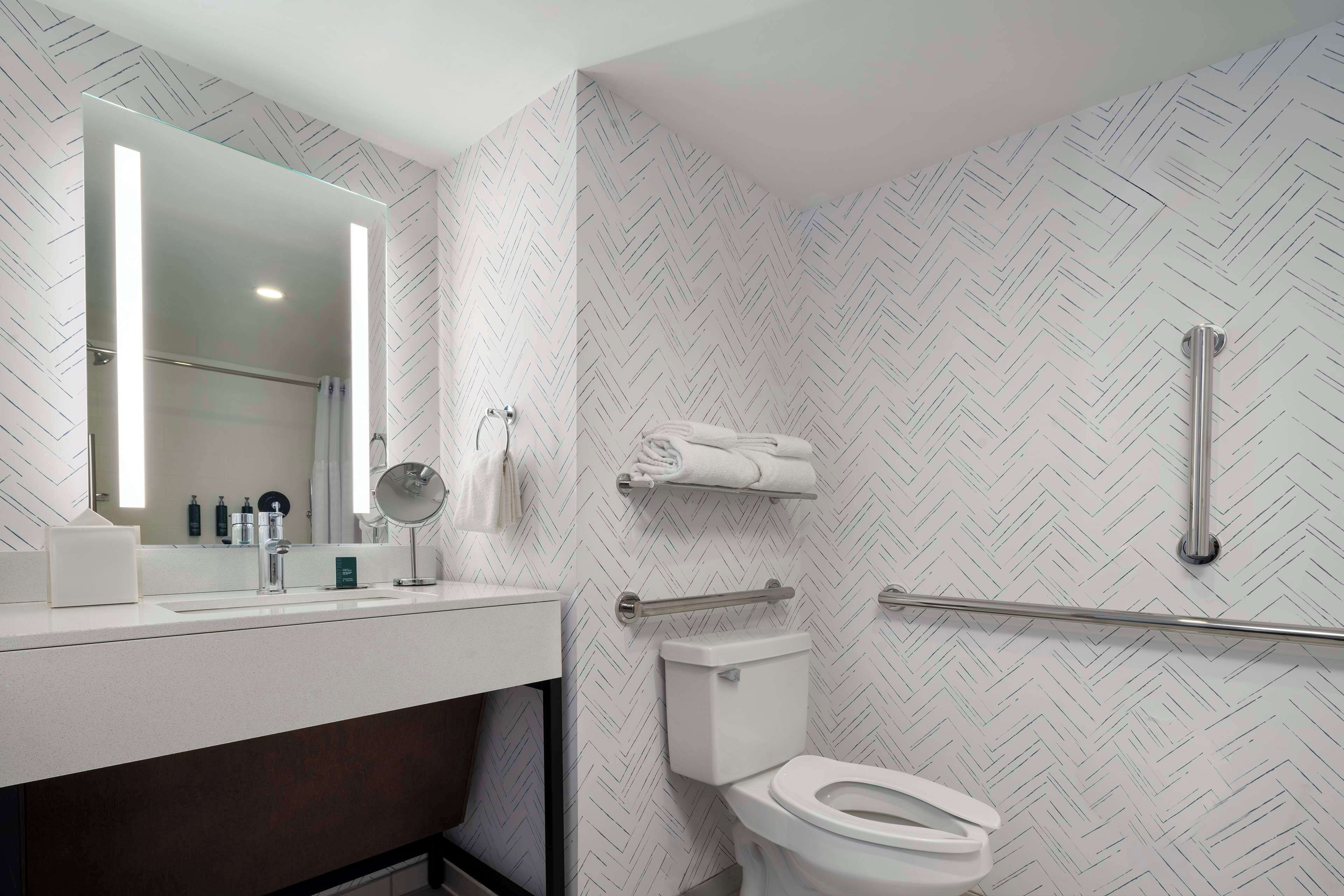 Un baño moderno con lavamanos, espejo y retrete blancos. Hay un perchero sobre el retrete y una zona de ducha con puerta de vidrio.