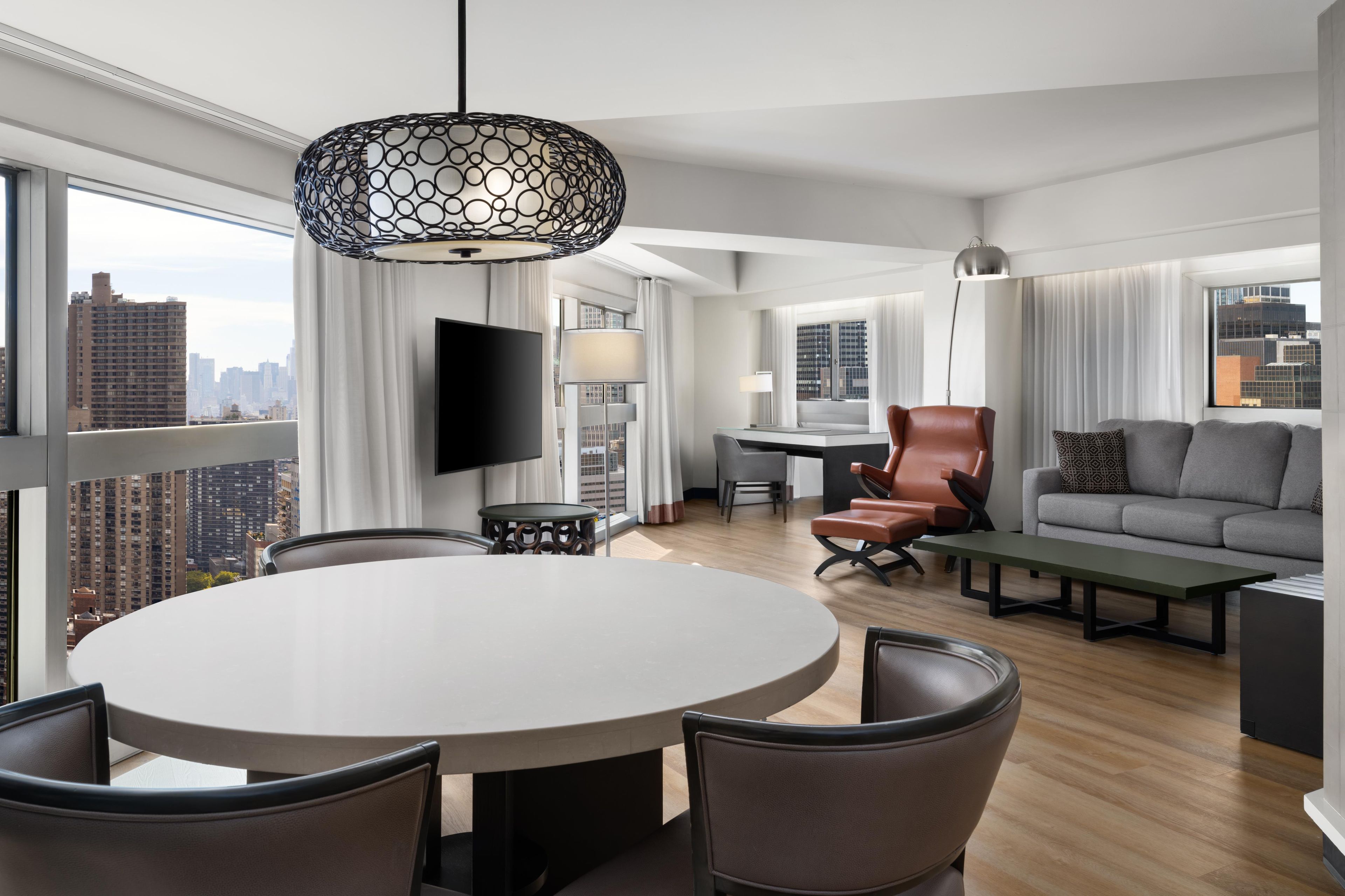 A modern hotel room with a round dining table, leather chairs, a sofa, a TV, and a city view.