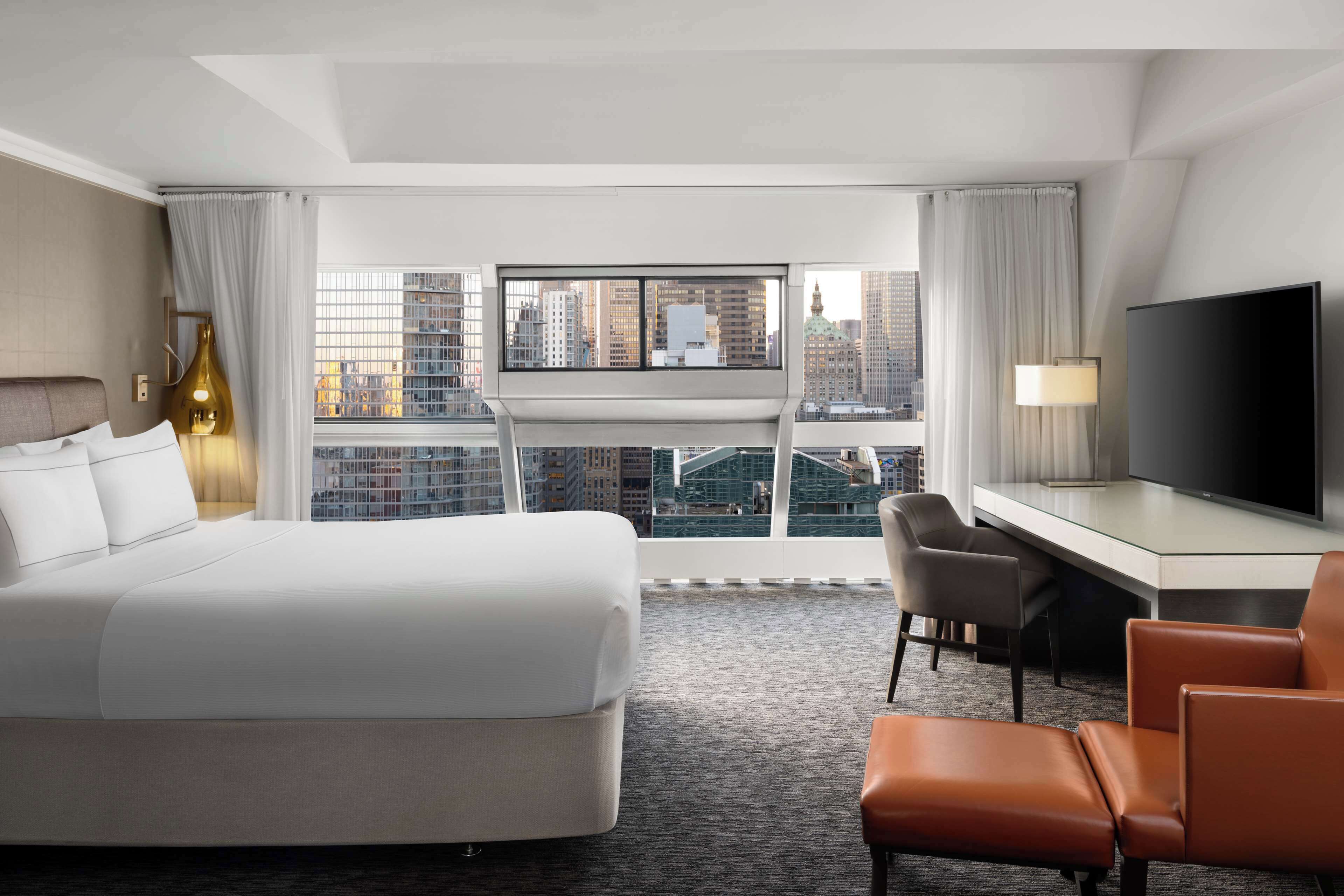 A modern hotel room with a large bed, a desk with a flat-screen TV, a chair, and a view of the cityscape through floor-to-ceiling windows.