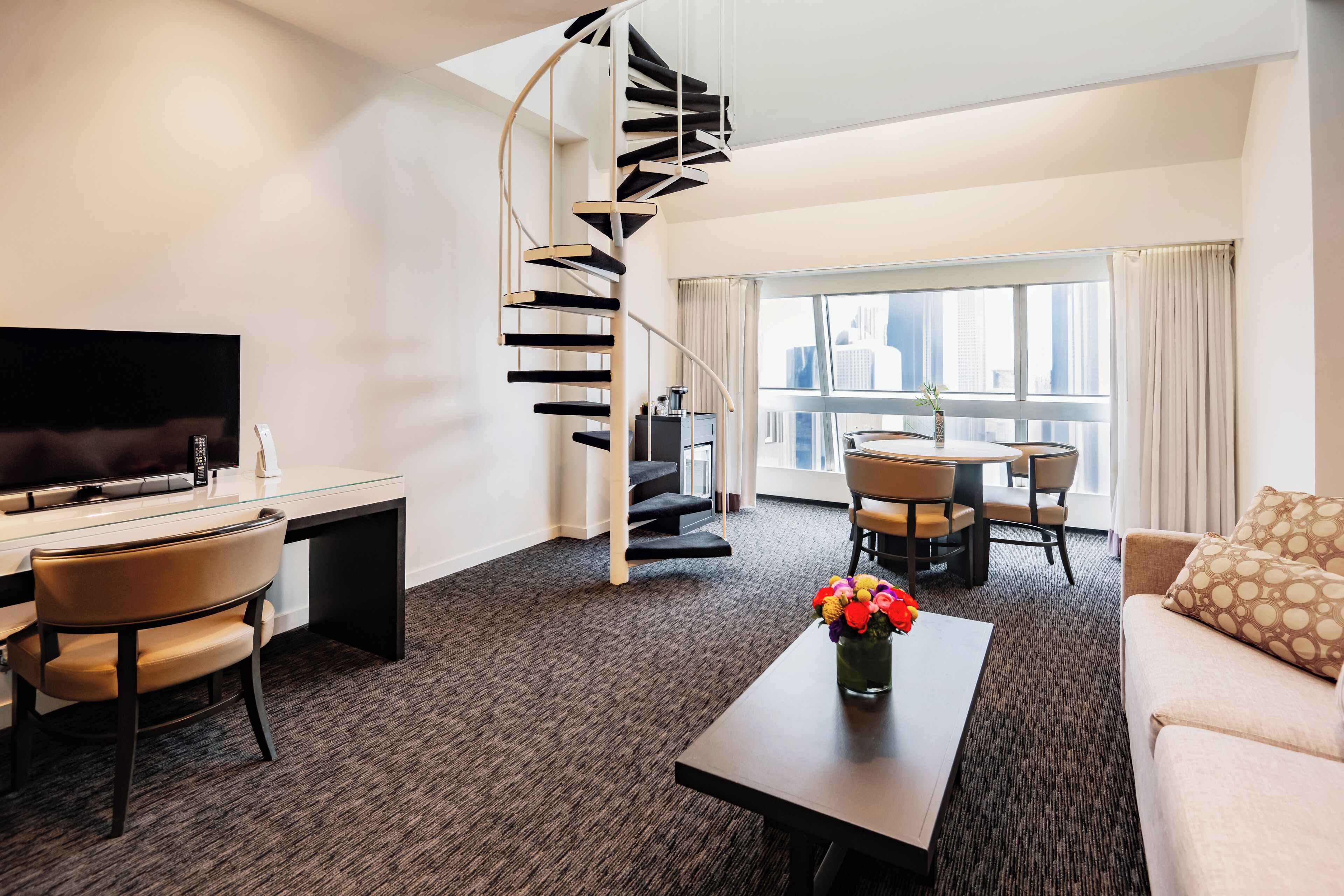 A modern hotel room with a spiral staircase, a flat-screen TV, a desk, a sofa, and a dining area with a view of the city.