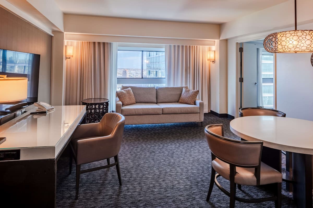 Une chambre d’hôtel avec un canapé, une table à manger, des chaises, un téléviseur et une grande fenêtre avec des rideaux.