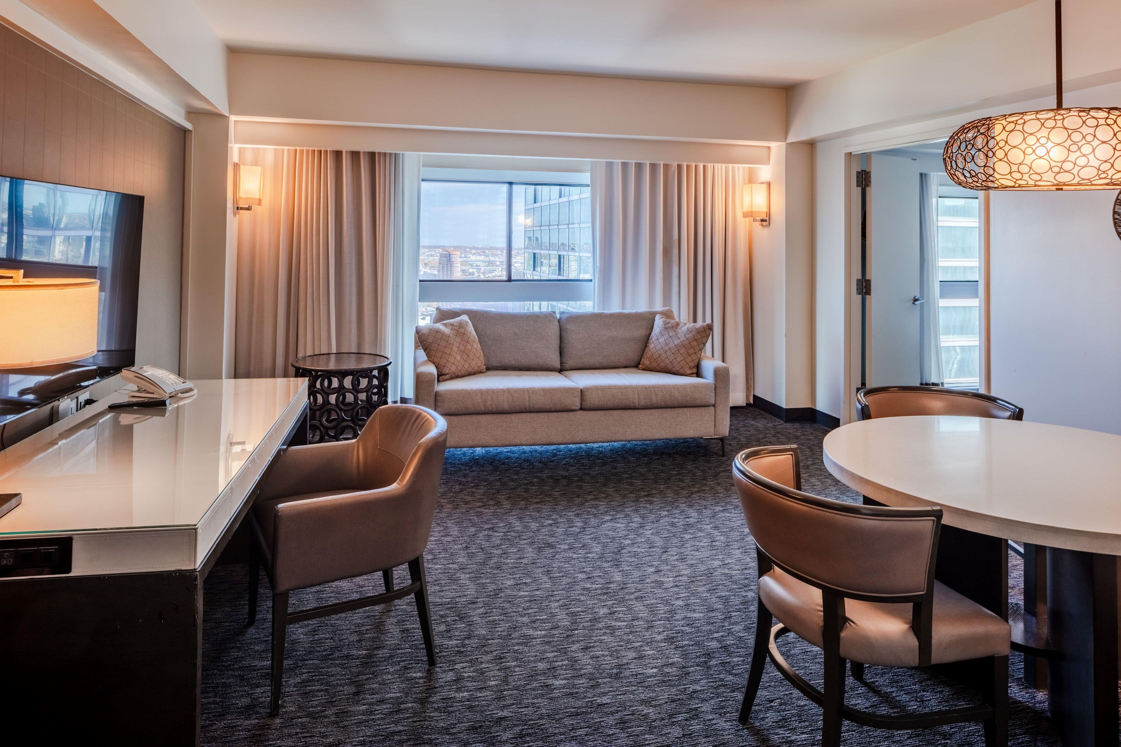Habitación de hotel con sofá, sillas, mesa de comedor, televisor y vista de la ciudad.