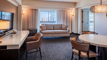 Habitación de hotel con sofá, sillas, mesa de comedor, televisor y vista de la ciudad.