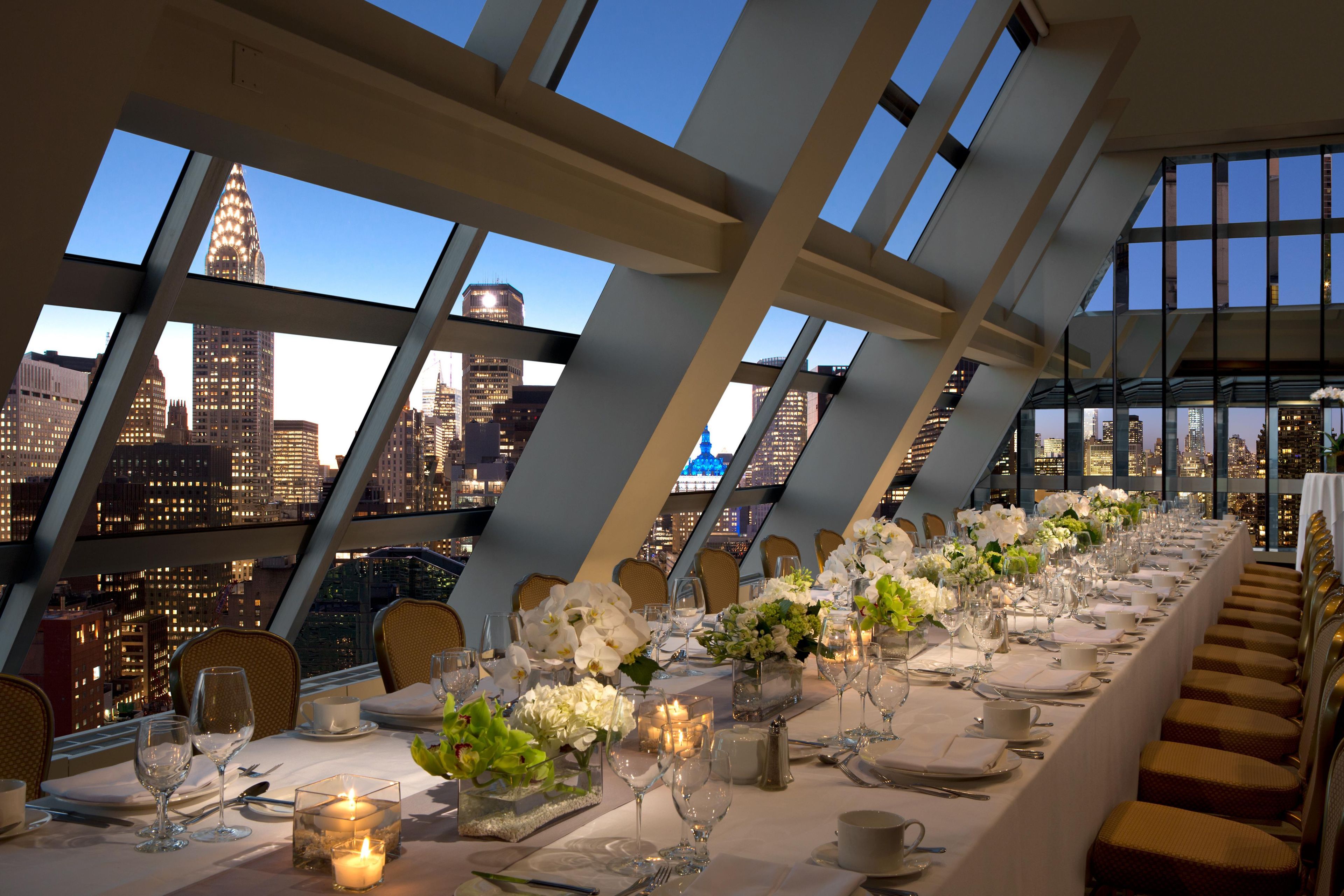 Una larga mesa de comedor lista para una comida, con vistas a un horizonte urbano a través de amplios ventanales.
