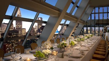 Una larga mesa de comedor lista para una comida, con vistas a un horizonte urbano a través de amplios ventanales.
