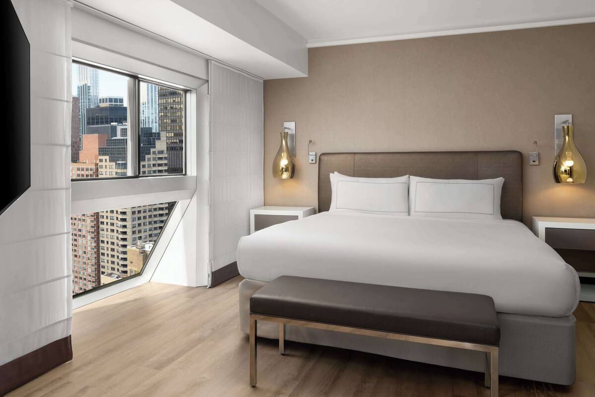 Une chambre d’hôtel moderne dotée d’un grand lit, d’une banquette, de deux tables de chevet et offrant une vue sur la ville grâce à de grandes fenêtres du sol au plafond.
