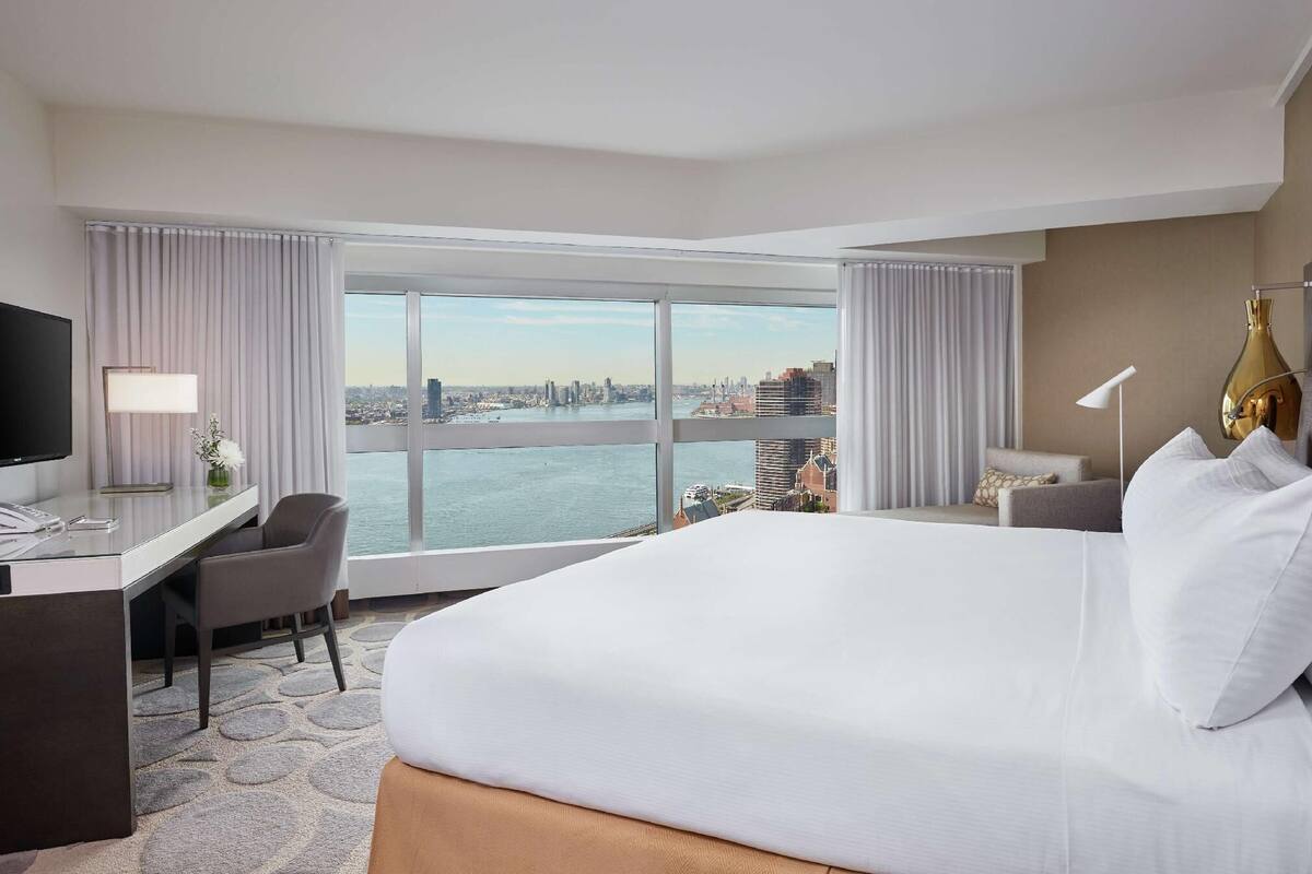 Ein Hotelzimmer mit einem großen Bett, einem Schreibtisch, einem Sessel, einem Fernseher und Blick auf eine Stadtlandschaft durch große Fenster.