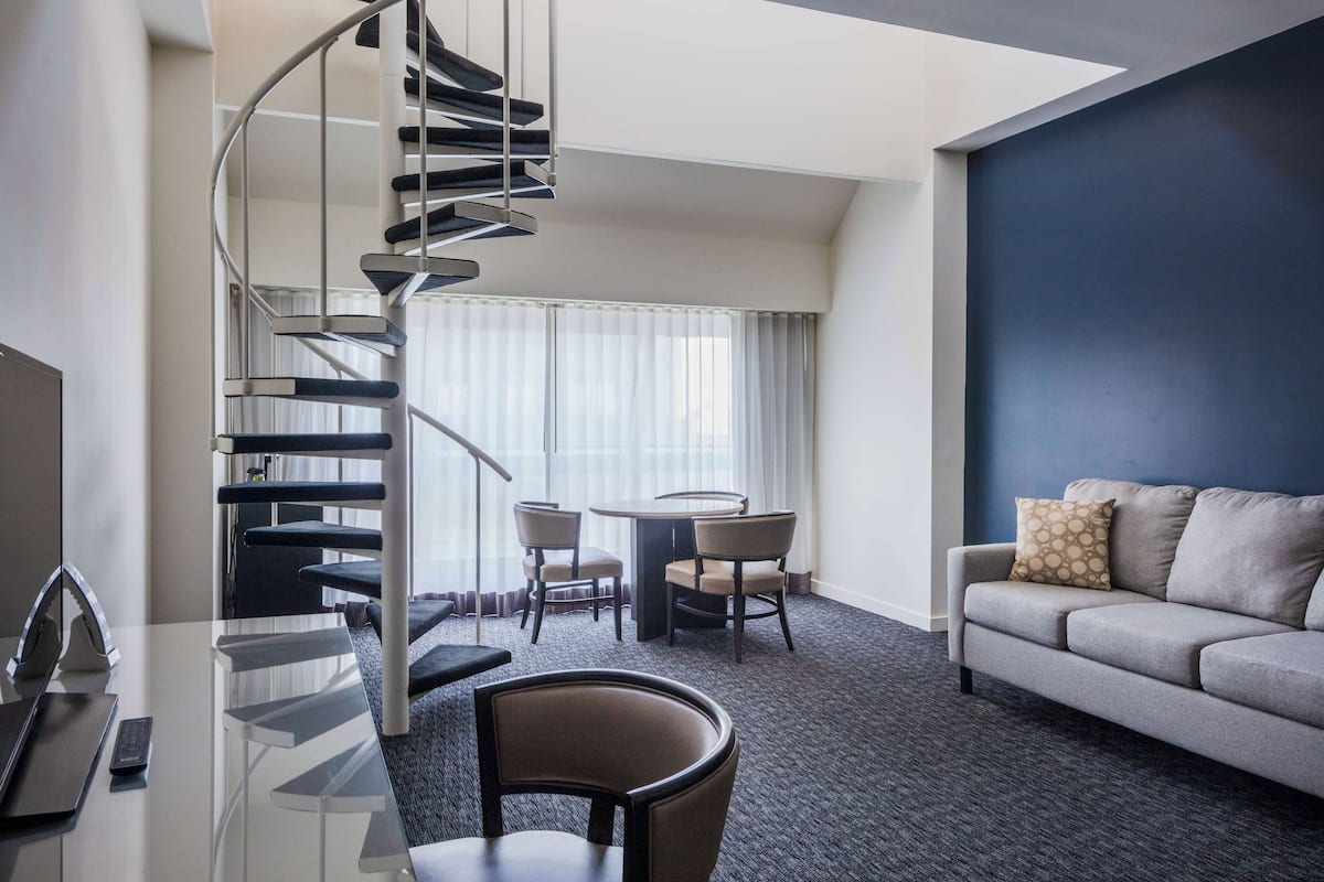 Une chambre d’hôtel moderne avec un escalier en colimaçon, un coin repas avec une table ronde et des chaises, un canapé agrémenté d’un coussin à motifs et une grande fenêtre avec des rideaux légers.