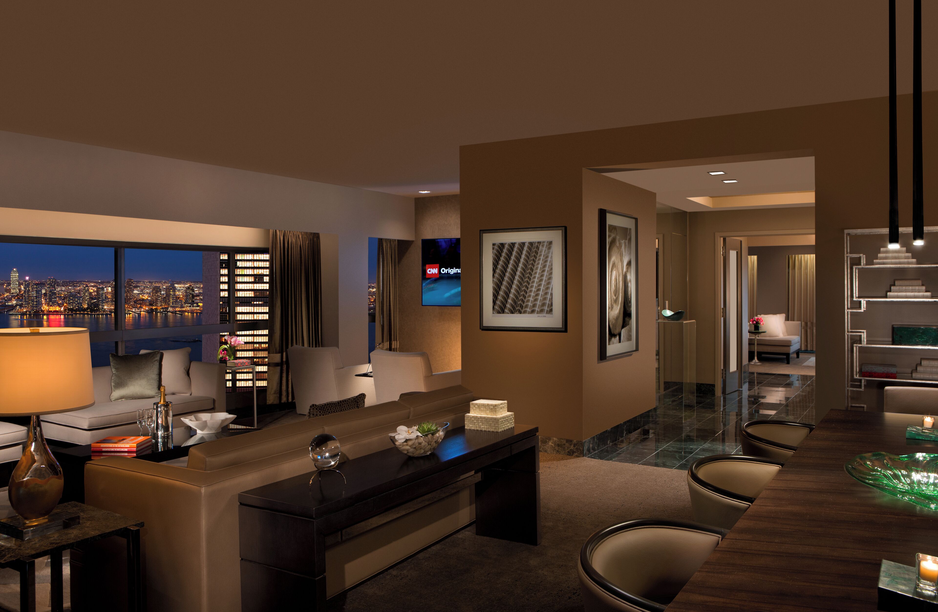 A modern hotel room with a city view, a desk with a lamp, a sofa, and a dining area with chairs.