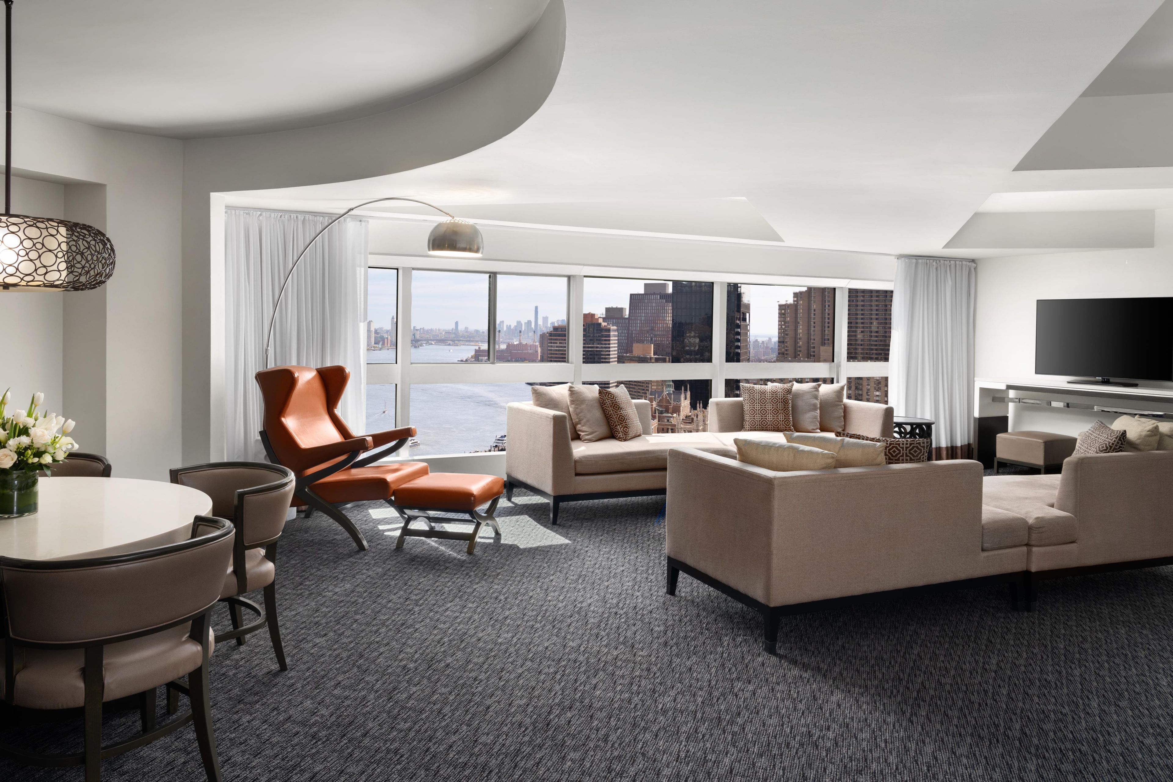 A modern hotel room with a city view, a dining area, and a living space with sofas and a TV.