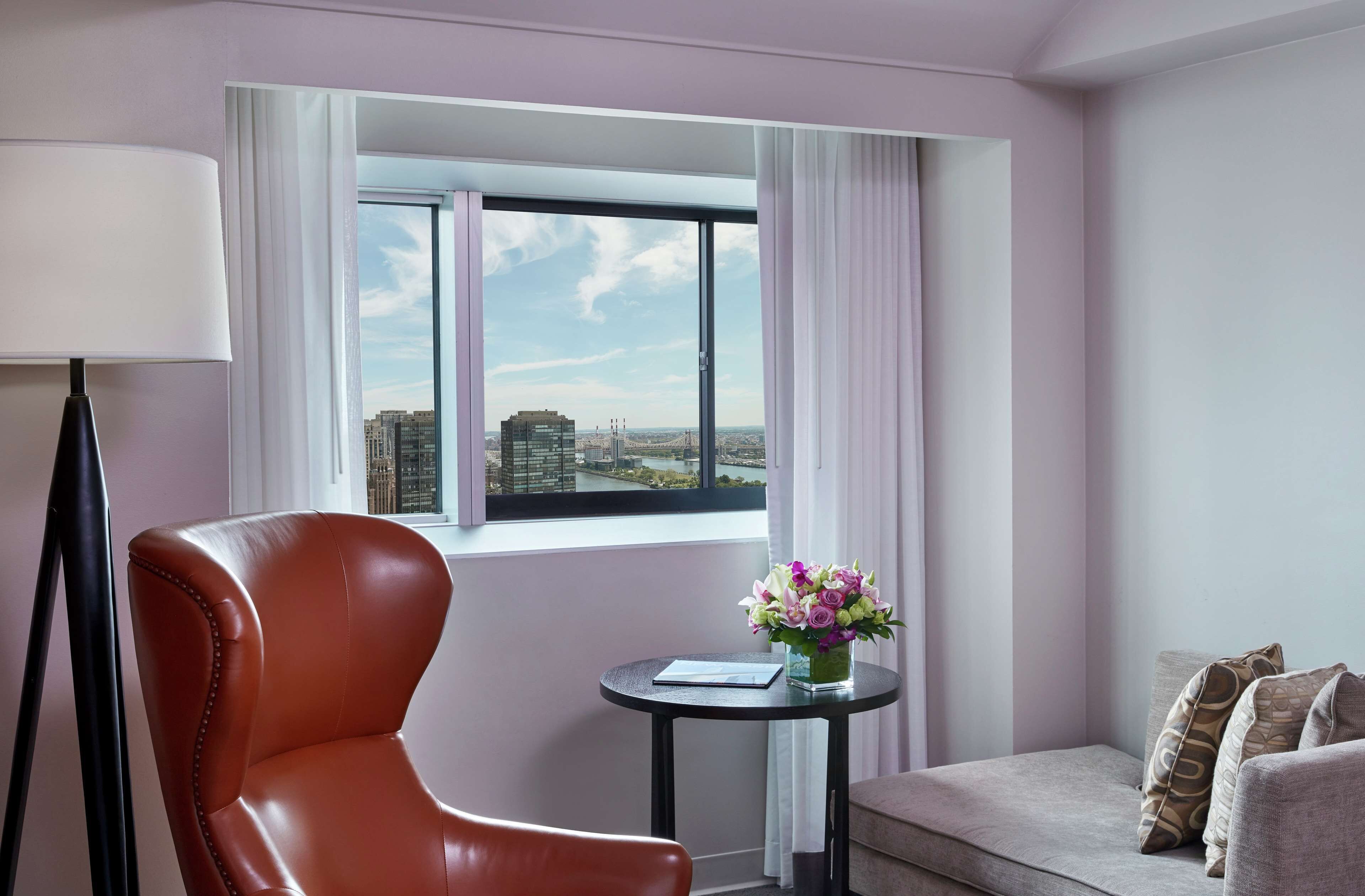 A hotel room with a large window, a leather chair, a small round table with a vase of flowers, and a sofa with patterned cushions.