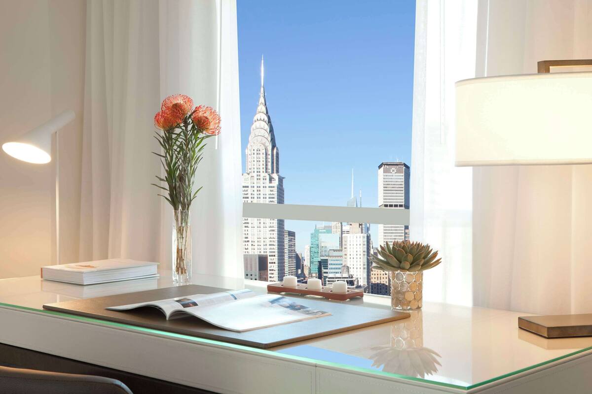 Une chambre d’hôtel moderne, équipée d’un bureau, d’un vase avec des fleurs, d’une lampe et offrant une vue sur le panorama urbain.