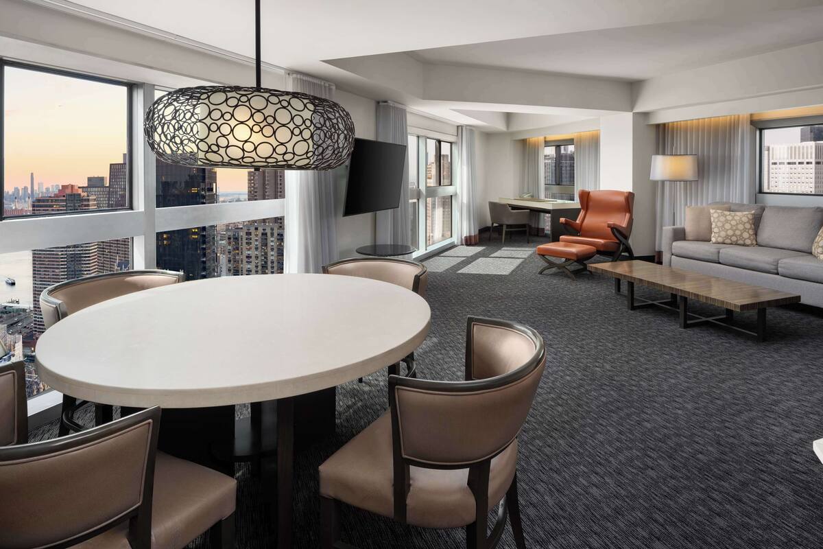 A modern hotel room with a city view, a round dining table, comfortable seating, and a large window.