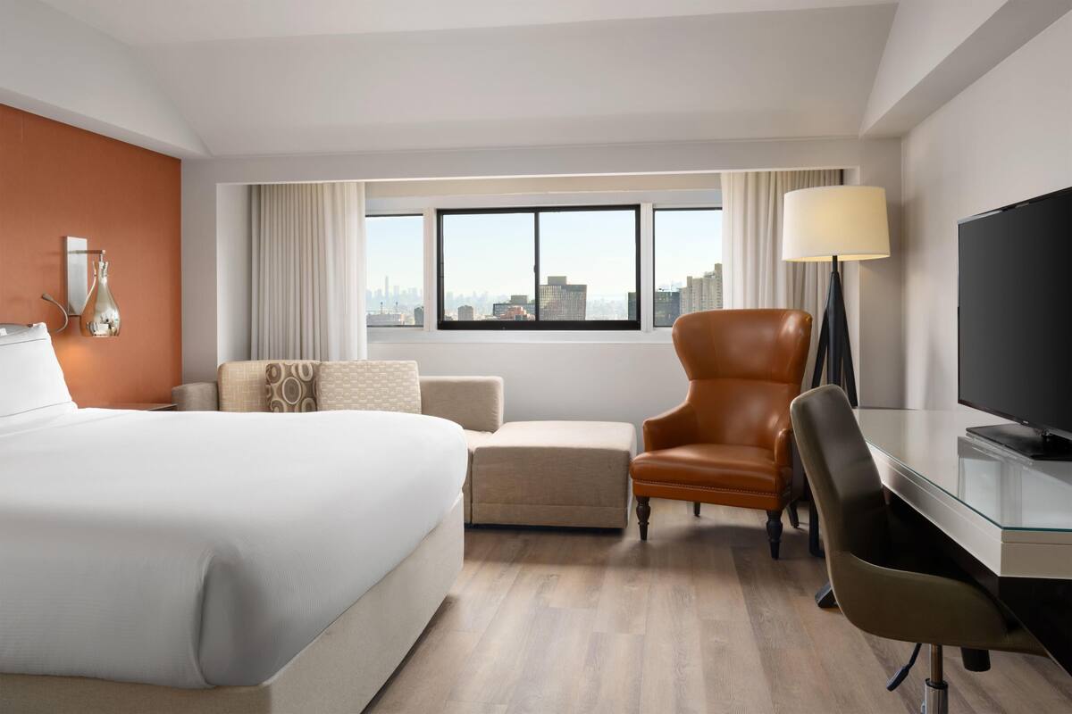 Une chambre d’hôtel comprenant un lit, un bureau avec une télévision, un fauteuil en cuir, un canapé et une grande fenêtre donnant sur la ville.