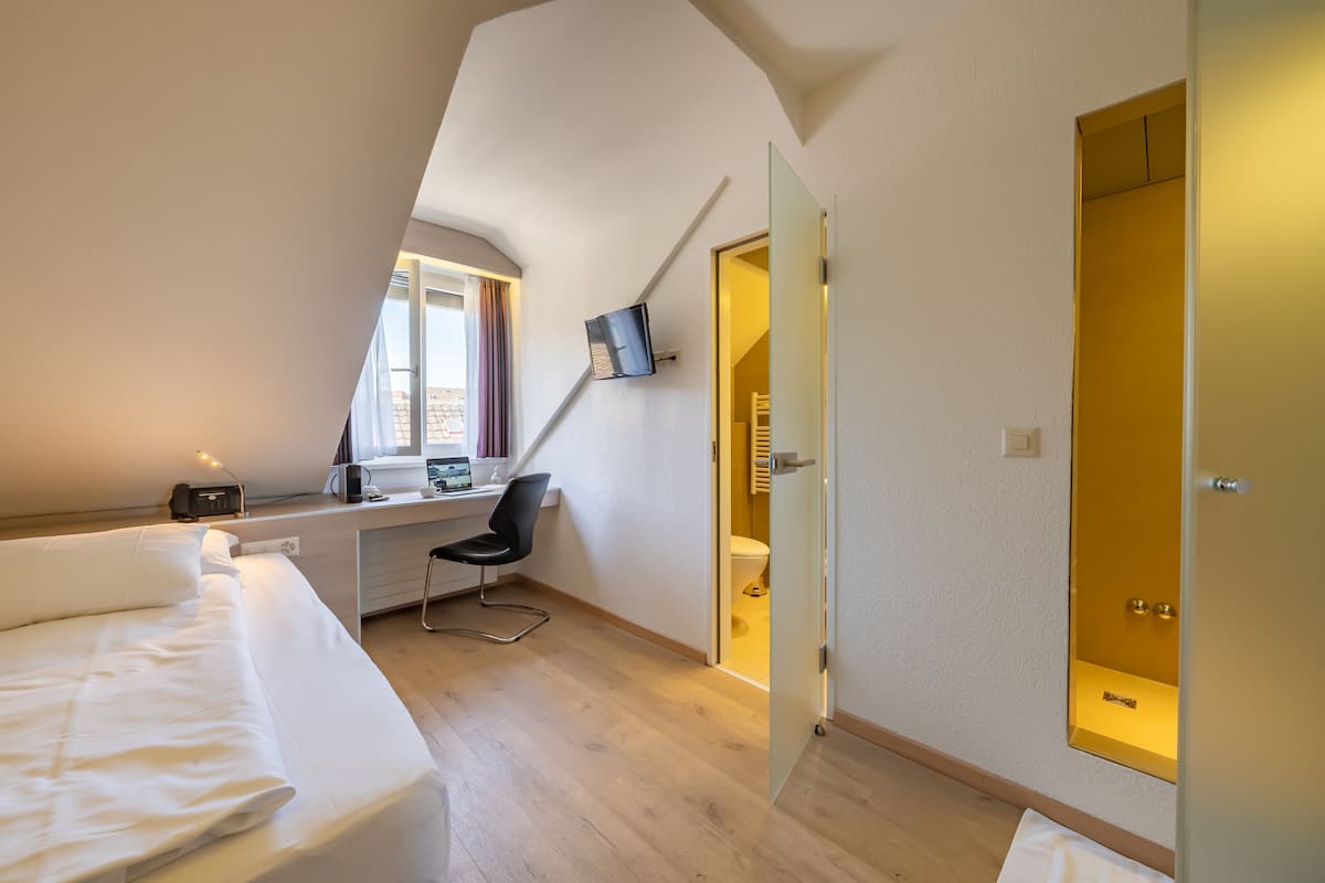 A modern bedroom with a bed, desk, and chair, a window with curtains, and a door leading to a bathroom.